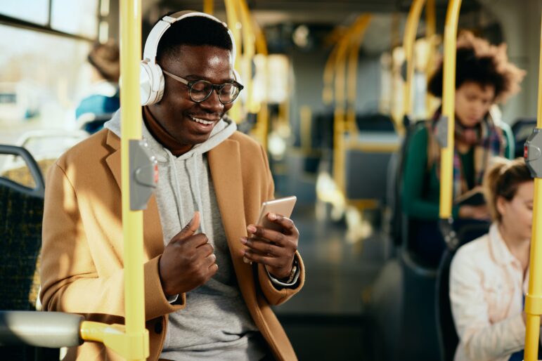 Person wearing headphones on public transit, smiling while using a smartphone, representing on-the-go audio learning for CME.